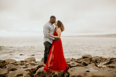 Woman in red dress hugs man as they stand on the rocks of La Jolla  during their California engagement session.