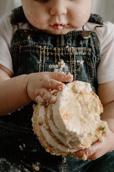 Milestone session cake smash of baby boy holding cake in his hands looking at it with cake on his face.