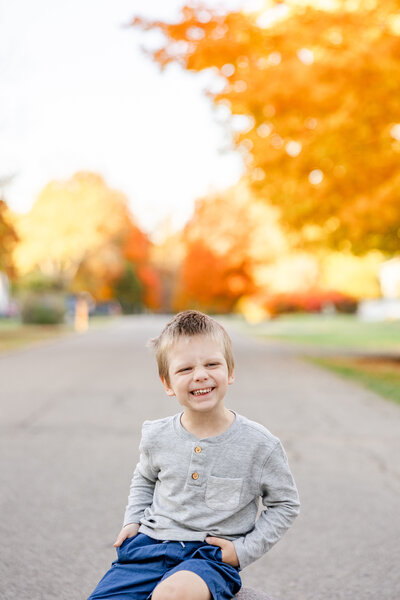 Young boy with hands in his pocket sitting on a stool in the street with beautiful orange Fall trees in the background.