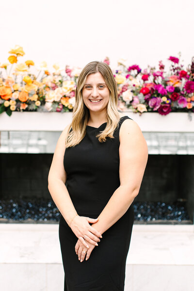 Kelsey Schultz, event sales coordinator at SoHo63, a Chandler wedding and event venue, smiles in front of a floral backdrop. Her approachable style and attention to detail help make every SoHo63 wedding and event seamless and memorable.
