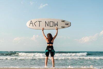 photo femme de dos sur une plage, portant une planche de surf au dessus de sa tête écrit "act now"
