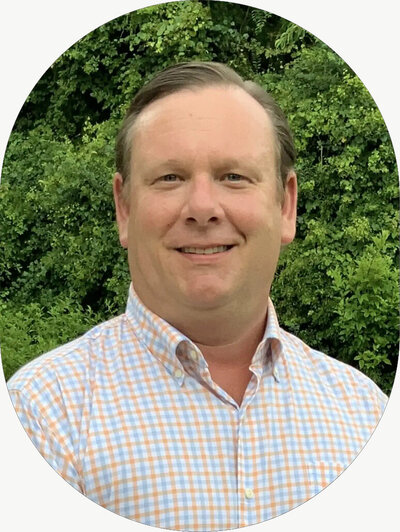A man standing outside in front of lush greenery, wearing a checked collared shirt.