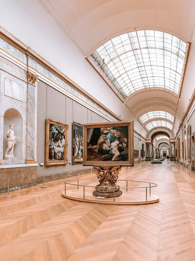 A spacious art gallery with a high arched ceiling and parquet wooden floors. Large framed paintings hang on the walls, with one prominently displayed on a stand in the center. A classical statue is visible to the left.
