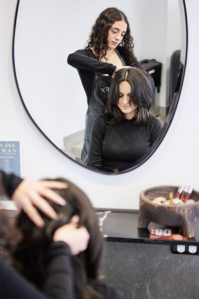 Hair specialist at Blake & Kwan Cronulla styling brunette hair during a silent haircut session, reflected in an oval mirror within a modern salon.