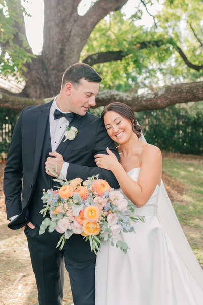 Bride and groom smiling together under a tree at One Main Eleven, holding a colorful pastel wedding bouquet.