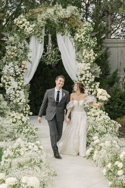 Bride and groom joyfully exiting their wedding ceremony adorned with an elaborate white floral arrangement at Chateaux at Fox Meadows
