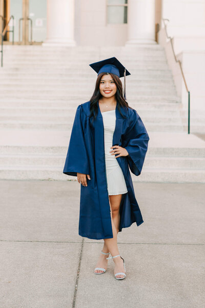 A high school senior in white dress and cap and gown stands downtown Albany, OR