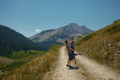 A destination wedding photographer sits cross-legged on a warm-toned studio backdrop, holding multiple vintage and modern cameras while smiling at a small dog. Surrounded by printed photos and old-school camera gear, she exudes a playful, nostalgic vibe.