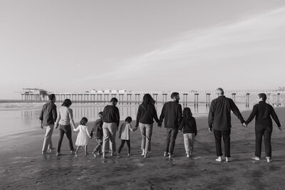 Black and white photo of a large family holding hands, facing the ocean near a pier, during a beach portrait session.