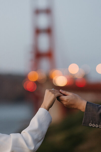 A close-up of a couple holding hands during a photoshoot, with the Golden Gate Bridge blurred in the background during twilight in San Francisco.