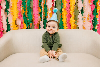 Toddler sits on a tan couch with a blue birthday hat on