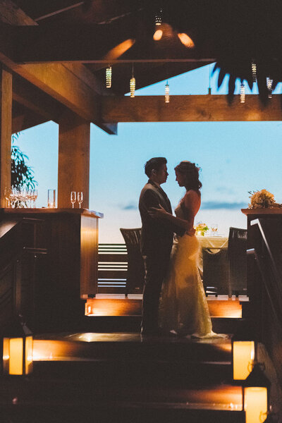 couple embraces on their wedding day under a barn structure with the sun setting behind them