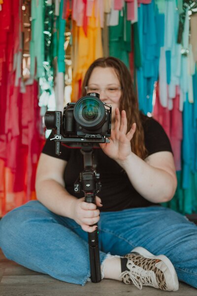 mara shields standing in front of the head table, with camera on a tripod, in a black dress and sneakers
