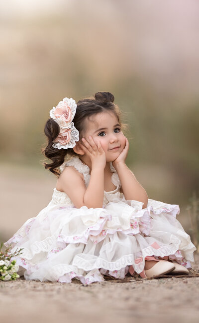 Young toddler girl wearing an off white lace vintage dress and a lace and peach bow on one side of her dark brown hair styled in pig tails sitting on a dirt path holding her face between her hands with her nude flat shoes showing from under her dress.