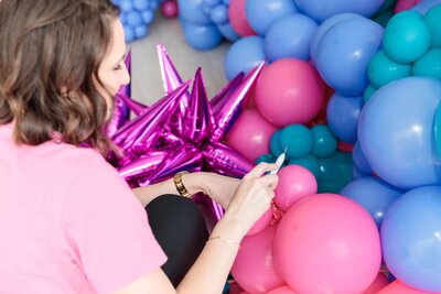 The expertise of Air with Flair, your go-to for premium balloons, decor, and rentals. The expert, dressed in a pink t-shirt and black pants, is crafting a stunning pink and blue balloon installation for the next unforgettable celebration.