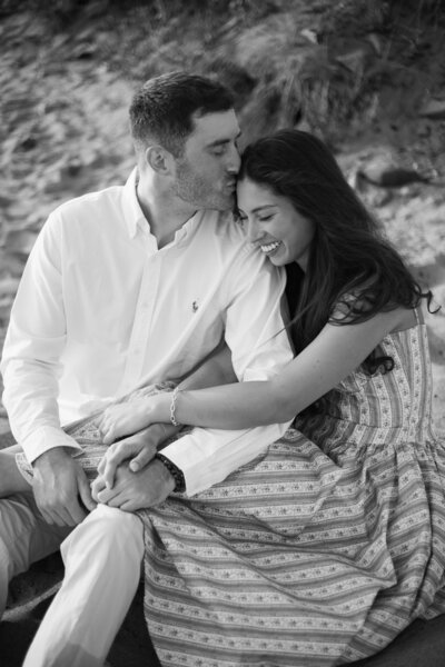 Collins Beach Newport RI | Kelsey Sheehan Photography Timeless Rhode Island Weddings | A couple sits on a beach with the man kissing the woman's head. She smiles warmly, wearing a patterned dress. The mood is affectionate and serene.