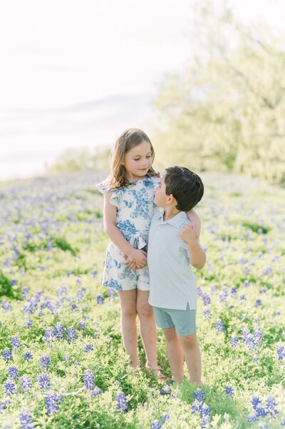 Brother and sister standing with arms wrapped around each other  and adoring smiles standing in a flower field.