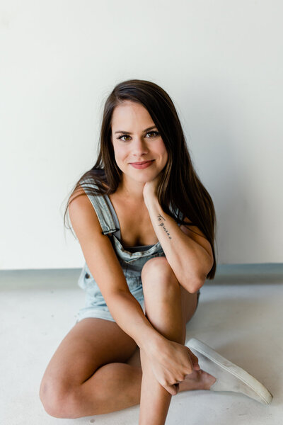 Girl with brown hair wearing blue overalls sitting on the floor in front of white wall leaning on her right hand, Charlotte boudoir photographer