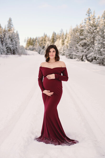 A pregnant woman wearing a red dress and short brown hair stands against a beautiful snowy winter background. Taken in 100 Mile House by Brittany Danielle.