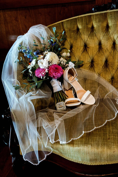 Bride's white wedding shoes paired with her veil and a bouquet of bright florals on a olive green vintage couch
