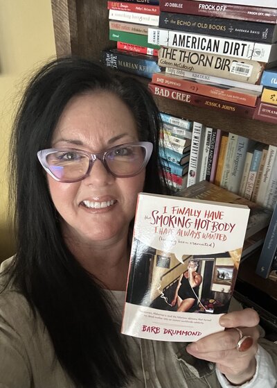 Barb Drummond standing in front of a bookshelf, holding her book "I finally have the smoking hot body I have always wanted (having been cremated)