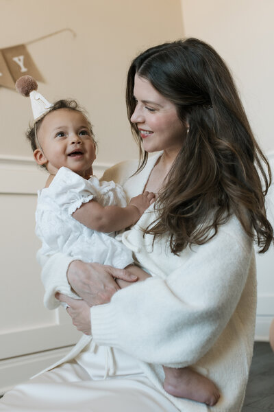 Mom is holding a baby girl for her first birthday smiling at her, they are both in cream colored outfits. 