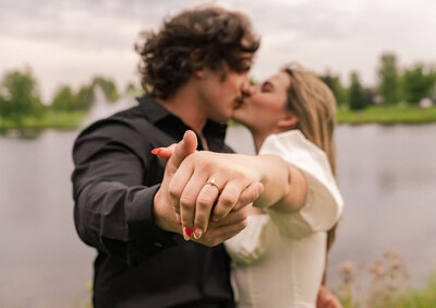 Close-up of an engaged couple sharing a kiss while holding out a hand to reveal the engagement ring, captured by Joseph Mayden in a romantic lakeside setting.