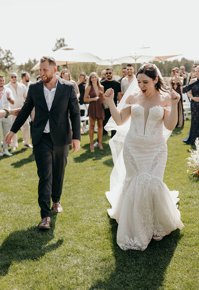 Bride and groom leaving their ceremony with joyful expressions, appearing to dance away together.