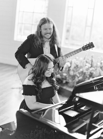 Black and white photo of Mikaela Kahn playing piano while her husband plays guitar