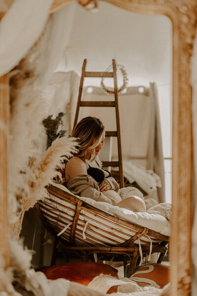 Femme enceinte, en sous-vêtements, assise dans grand fauteuil rond en bois avec coussin dans un décor cosy, à mon studio photo en Vendée.