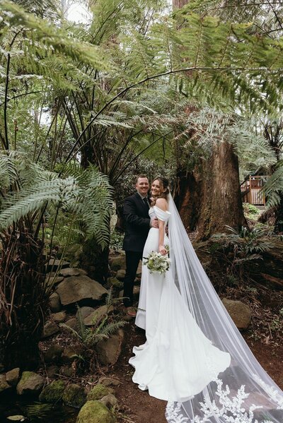 A bride with a white dress and long veil hugging her groom and standing in a forest.