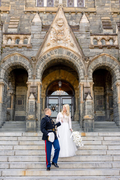 A bride in a white dress and a groom in a formal military uniform stand on stone steps in front of an ornate, gothic-style building. The bride holds a bouquet of white flowers as they lovingly gaze at each other.