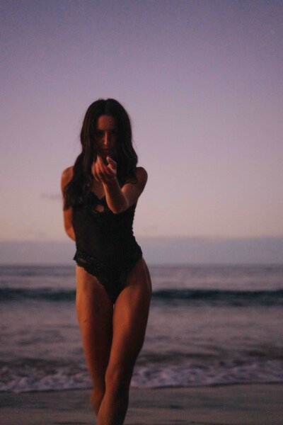 Meg O'Neill walking away on the beach with a black swimsuit surrounded by water and sky