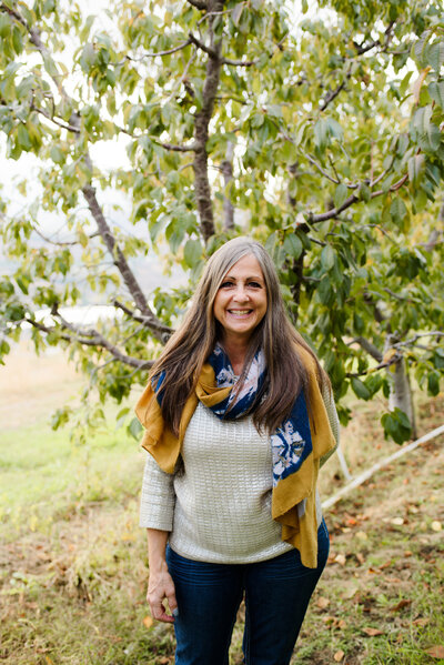 A smiling woman standing next to an apple tree in East Wenatchee. She is wearing a light colored sweater with a blue and yellow scarf and blue jeans.