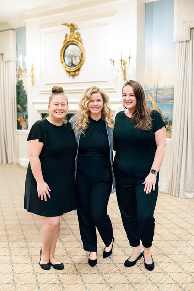 Three women wearing black outfits stand together, smiling, in a room with elegant decor. A mirror and chandeliers are visible in the background, and the carpet is patterned. The atmosphere appears formal and friendly.