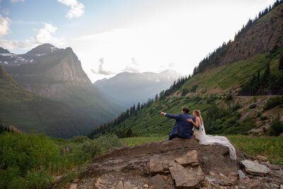 A bride wearing a white dress sits on a rock with her groom as he points out to something in the distance.