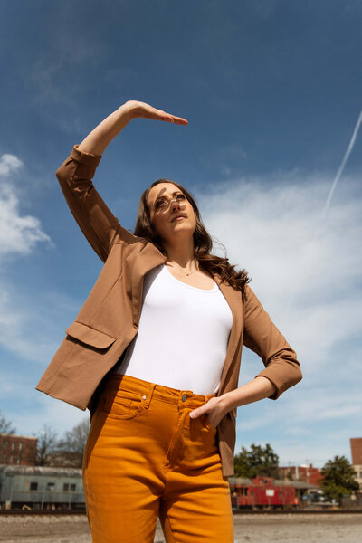 A woman stands outdoors under a blue sky with scattered clouds, wearing a brown blazer, white top, and mustard yellow pants. She poses with one arm curved above her head and looks upward. Buildings and trees are in the background. 