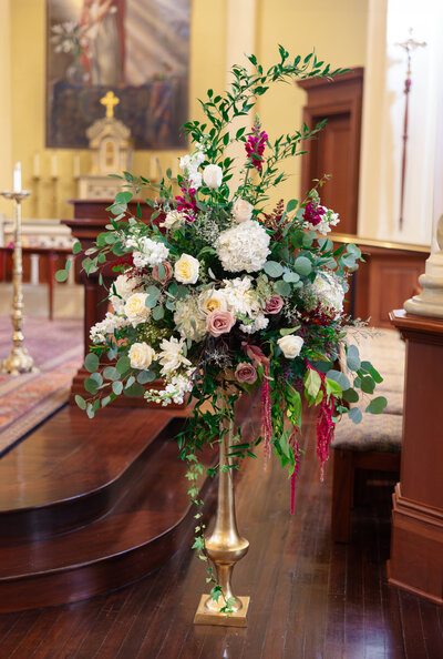 A tall, elegant floral arrangement in a church features a mix of white, pink, and burgundy flowers with lush green foliage in a golden vase. The background shows a church altar with religious decor.