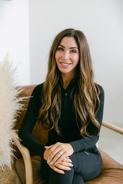 A woman with blonde hair wearing black scrubs sits smiling on a brown chair in a bright room at Falmouth Maine Medical Aesthetics, with pampas grass visible to the side.