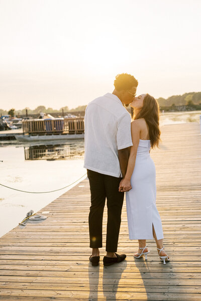 Engaged couple kissing on dock by the water at sunset
