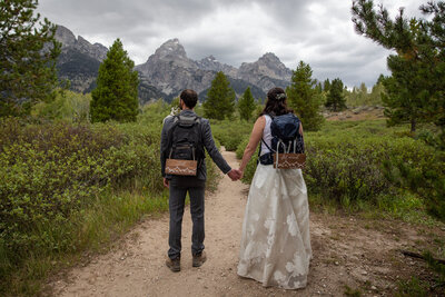 Bride and groom hold hands and face down the trail towards towering mountains in the distance,  they have hand-painted signs hangin from their backpacks that read "Just" & "Married"