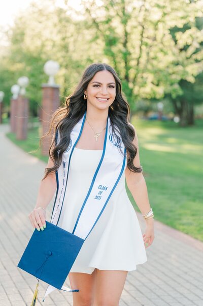 A woman walks and smiles in a white dress holding her graduation cap at Quinnipiac. 
