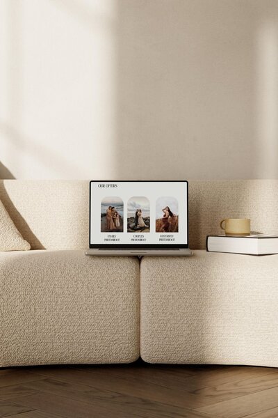 Open laptop on cream boucle sofa showing a photographer’s website offers for family, couples and maternity photoshoots, with coffee cup and book beside it.