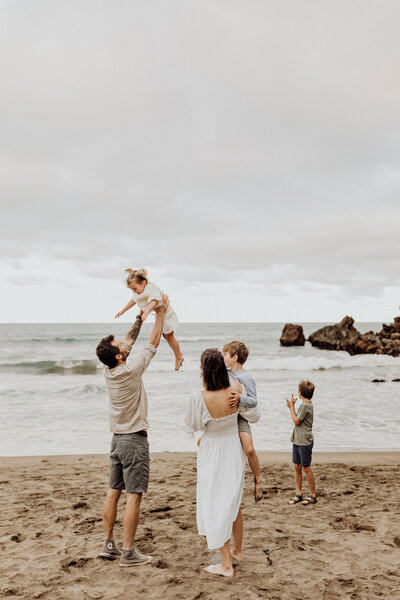 A father throws his baby in the air while the mother carries the young son and the older brother is looking out at the ocean in a candid family photograph taken by christchurch photographer lucy jane