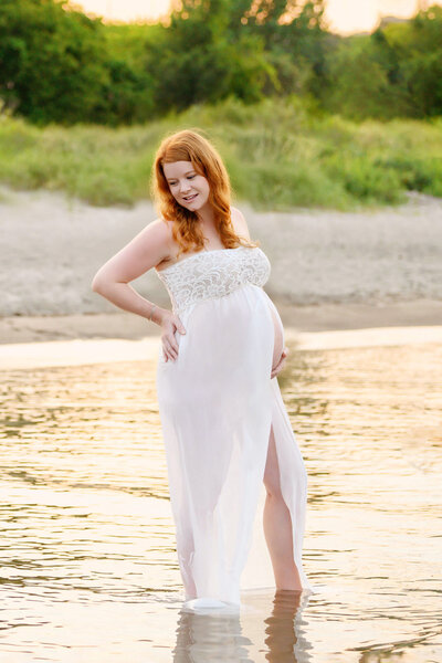 Red-haired mom wearing a white maternity dress at the beach at sunset for her outdoor beach maternity photography session in Burlington, Ontario