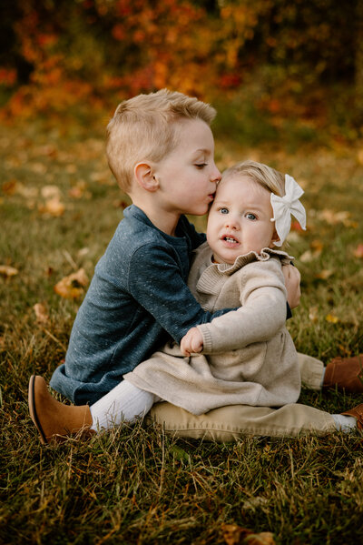 Toddler boy sitting on grass with baby sister sitting on his lap while hes kissing her temple with beautiful fall trees behind them in Crofton Maryland.
