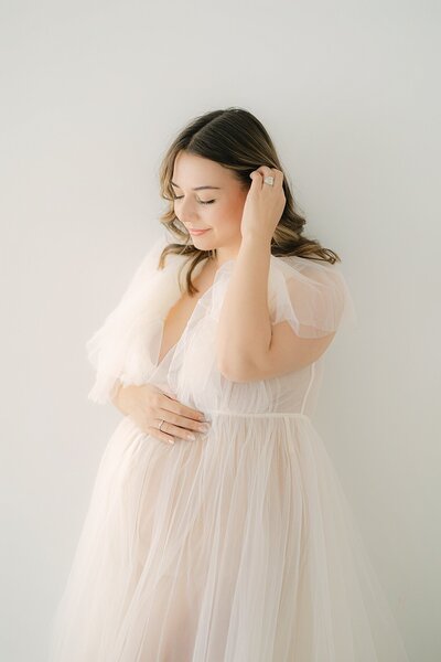 Pregnant mother in cream tulle dress tucks her hair behind her ear during session with Indianapolis maternity portrait photographer