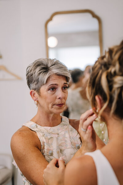 Mom wiping tears from daughters face as she puts on wedding dress.