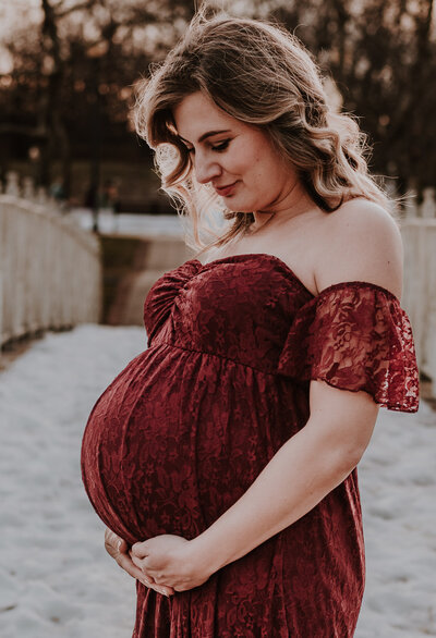 Pregnant mom standing on snowy bridge in Annapolis Maryland wearing a red dress and looking down at her pregnant belly while holding it in Annapolis Maryland.