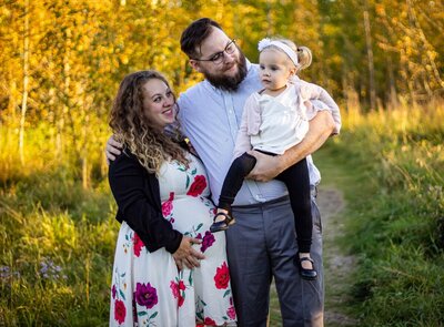 a pregnant woman smiles at her daughter who is on her dad's arms